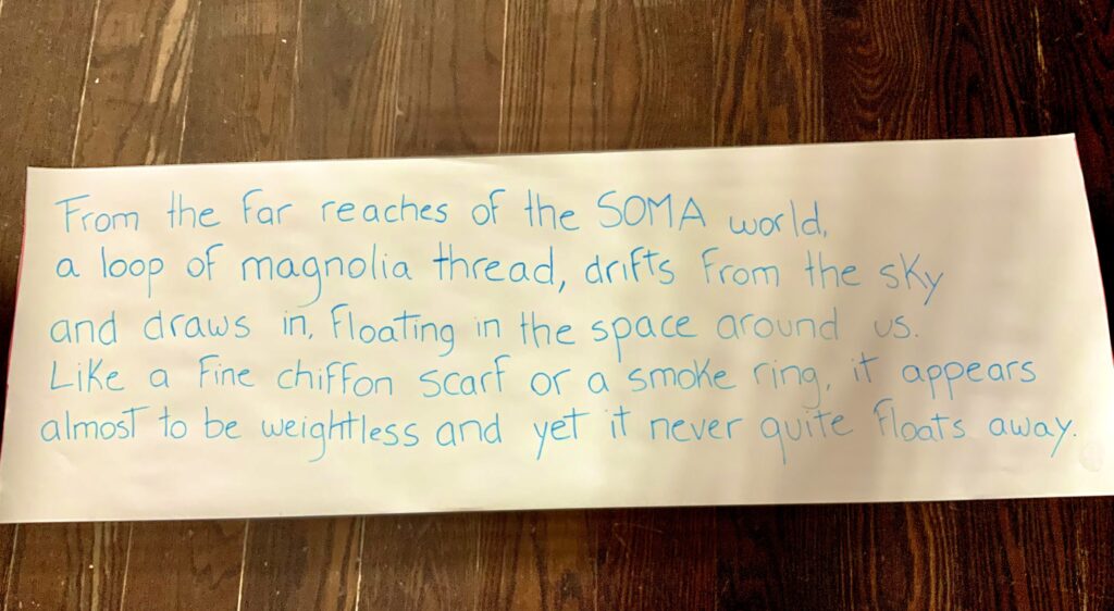 A colour photo of a large piece of paper laid out on a wooden floor with blue hand written text. It reads: 'from the far reaches of the SOMA world, a loop of magnolia thread, drifts from the sky and draws in, floating in the space around us. Like a fine chiffon scarf or a smoke ring, it appears almost to be weightless and yet it never quite floats away.'