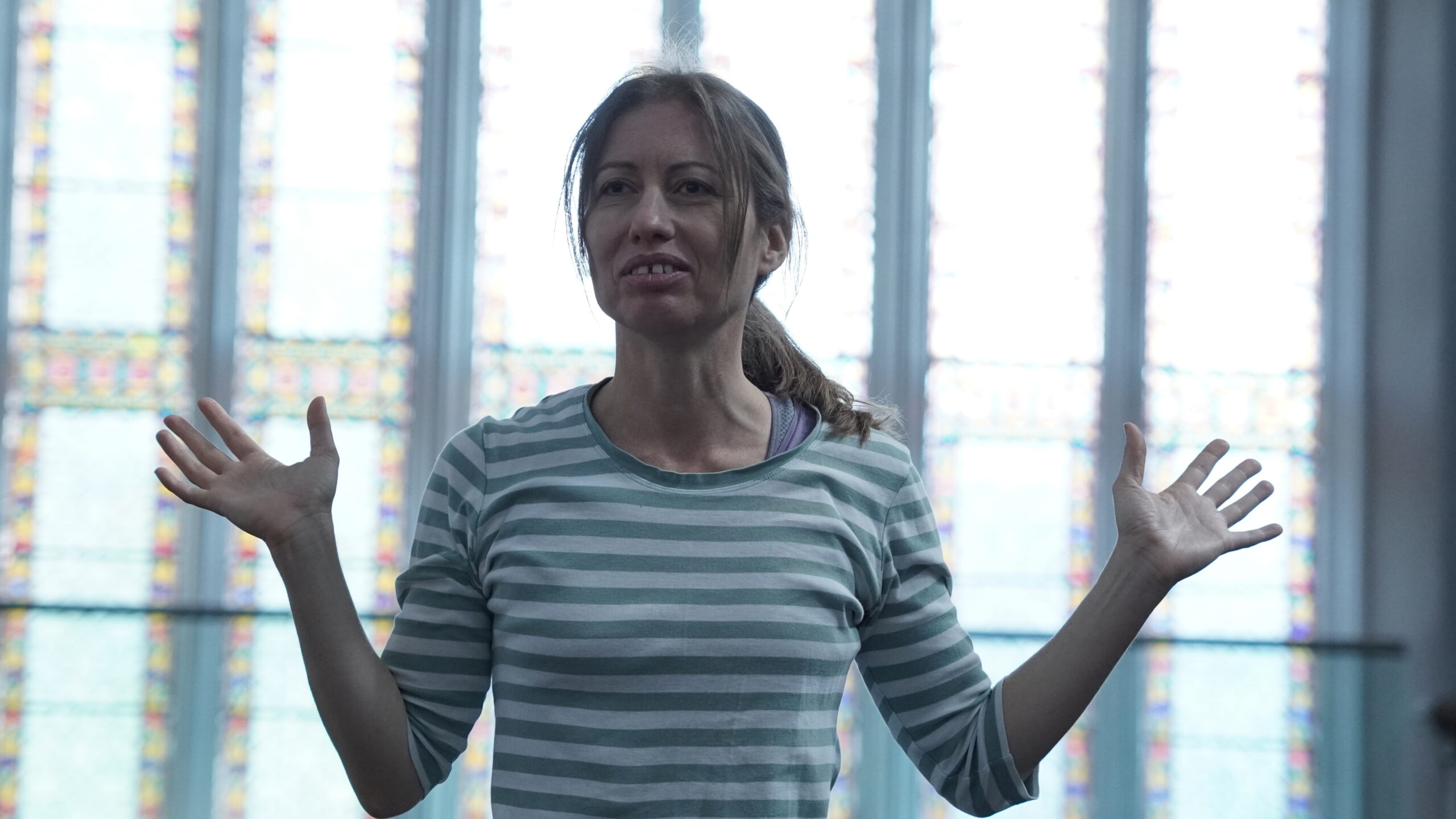 Holly, a fair skinned woman, is pictured front on and from the waist up, arms wide and palm of hands up in an expressive gesture of openness. She is wearing a green and white striped sweatshirt and has her hair in a ponytail. She is smiling. Behind her in soft focus is a large floor to ceiling window with oblong panes of stained glass.